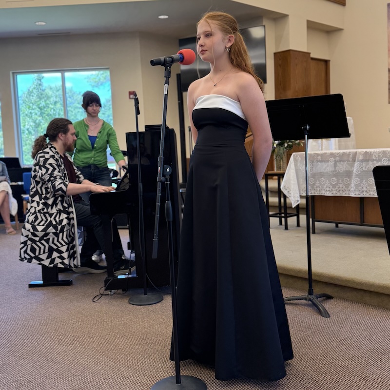 Student vocalist performing at a microphone during a recital with piano accompaniment in the background.